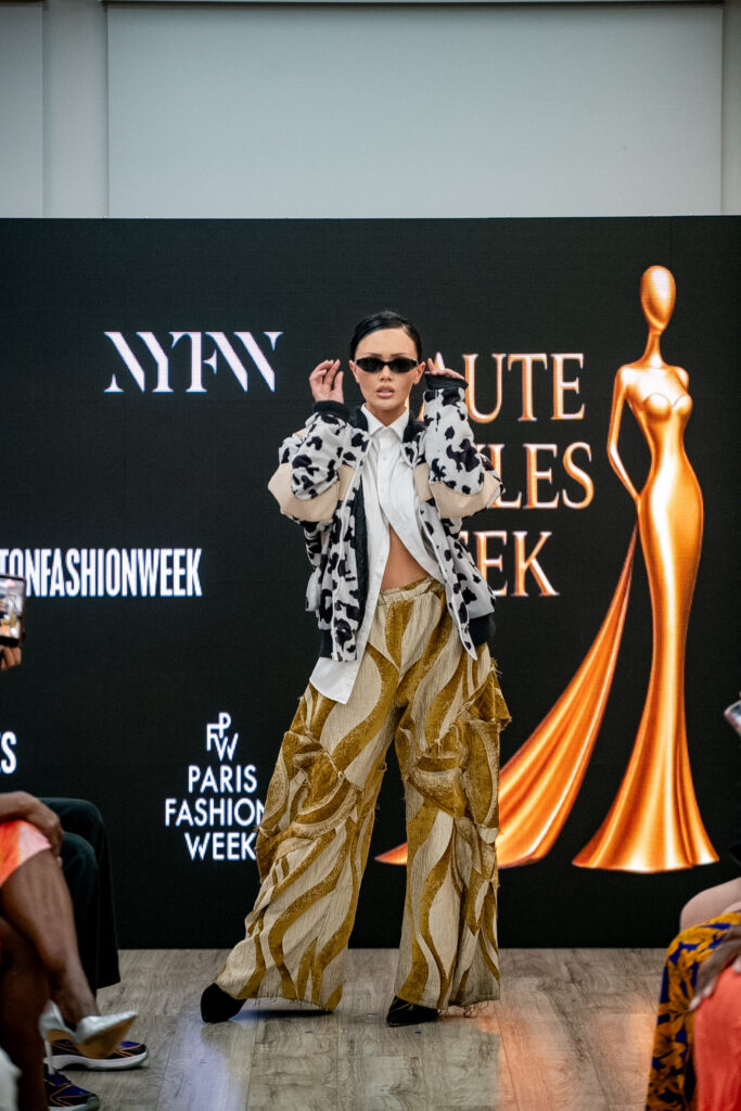 Model in a black and white fleece jacket and gold patterned trousers posing on a NYFW runway.
