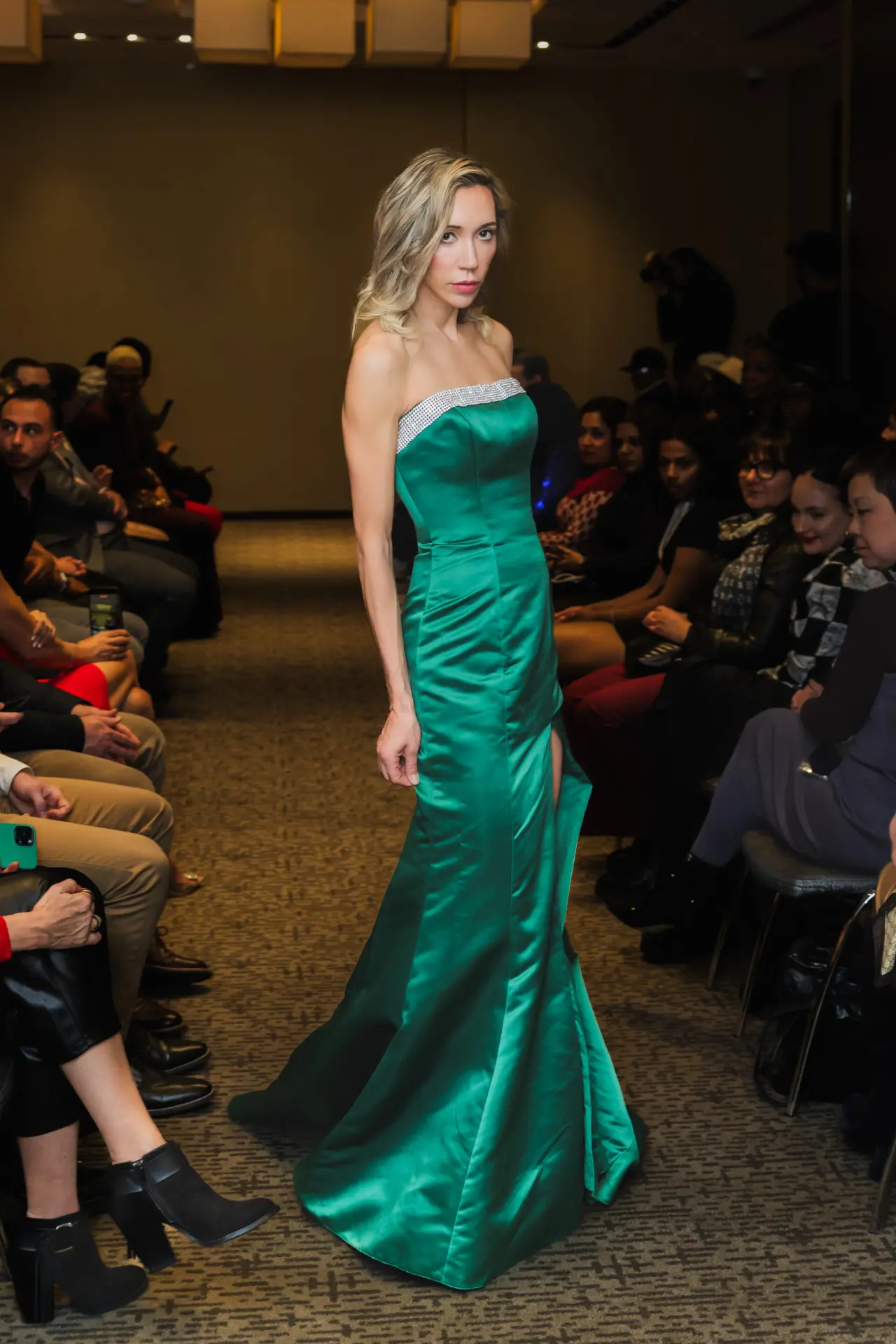 Model in an emerald green satin gown with a rhinestone neckline walking an indoor runway.